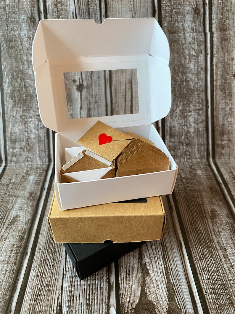 May include: A set of small brown envelopes with red hearts on the front, stacked inside a white box with a clear window. The box is sitting on top of a brown box and a black box.