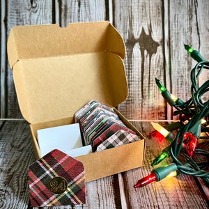 May include: A brown cardboard box with a hinged lid is open, revealing a set of 24 small envelopes with plaid and floral patterns. The envelopes are stacked inside the box. A single envelope with a red and black plaid pattern is sitting outside the box, with the number "24" in gold lettering on it. A strand of green Christmas lights with red and yellow bulbs is in the background.