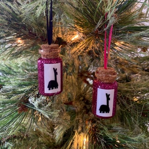 May include: Two pink glitter filled glass bottles with cork stoppers and black llama silhouettes on white labels. The bottles are hanging from a Christmas tree with black and red ribbon.