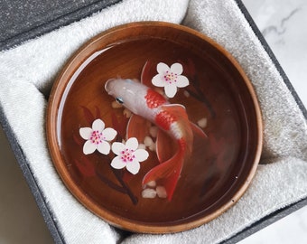May include: A small wooden bowl with a clear resin top containing a white and red goldfish with pink cherry blossoms. The bowl is in a black box with a white lining.