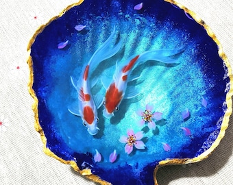 May include: Two red and white goldfish swim in a blue resin shell with pink flower accents. The shell is painted with gold trim.