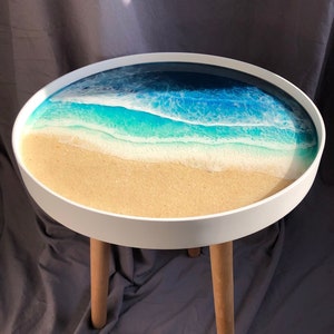 May include: A round white table with wooden legs features a resin tabletop with a blue and green ocean wave design and a sandy beach.