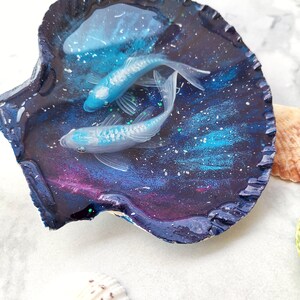 May include: A seashell dish with a galaxy-themed resin design featuring two white fish swimming in a blue and purple galaxy. The dish is on a white surface.