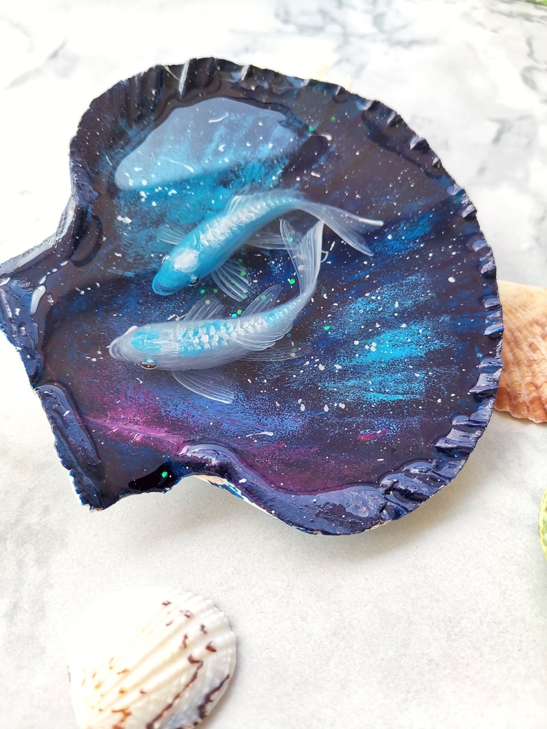 May include: A seashell dish with a galaxy-themed resin design featuring two white fish swimming in a blue and purple galaxy. The dish is on a white surface.