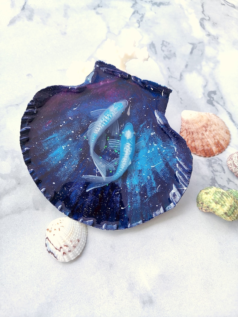 May include: A blue and purple seashell with a galaxy design, filled with two white resin fish. The seashell is on a white marble surface with other seashells.