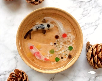 May include: A wooden bowl with a clear resin insert containing three white and black fish, white pebbles, and green leaves. The bowl is on a white marble surface.