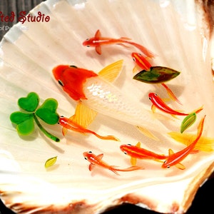 Puede incluir: Una concha decorativa con una escena pintada de peces koi y un tr&eacute;bol. Los peces son naranjas y blancos, nadando en resina transparente. La concha es blanca con bordes marrones. The Gifted Studio.