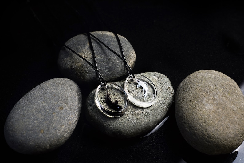 May include: Two black cord necklaces with circular pendants featuring a black and white fish design. The necklaces are displayed on gray stones.