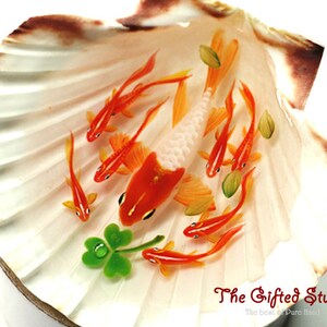 Puede incluir: Una pieza de arte decorativa de concha marina con peces koi naranjas y blancos en miniatura nadando en una concha. La concha es blanca con bordes marrones, y tambi&eacute;n hay un tr&eacute;bol verde de cuatro hojas. El texto "The Gifted Studio" es visible.