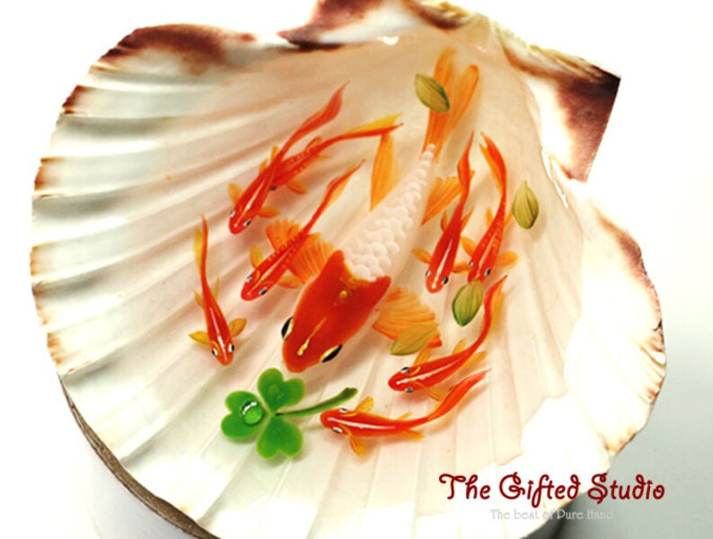 Puede incluir: Una pieza de arte decorativa de concha marina con peces koi naranjas y blancos en miniatura nadando en una concha. La concha es blanca con bordes marrones, y tambi&eacute;n hay un tr&eacute;bol verde de cuatro hojas. El texto "The Gifted Studio" es visible.