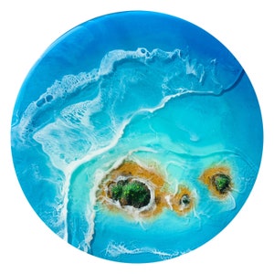 May include: Abstract resin art painting depicting a blue ocean with white waves and two green islands. The islands are surrounded by a golden sand beach.