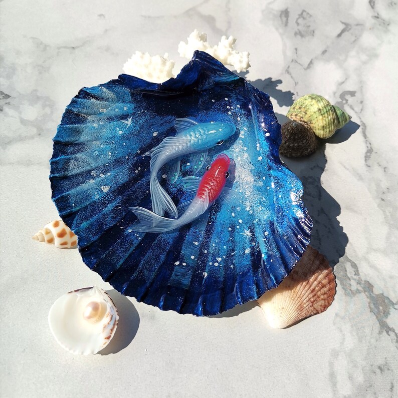May include: A blue and white seashell with a painted design of two fish, one red and one blue, on a white background.