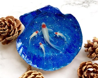 May include: A blue seashell with a white and red goldfish painted inside. The seashell is on a white background.