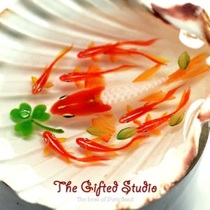 Puede incluir: Una concha decorativa con peces dorados en miniatura de color naranja y blanco nadando en su interior. Tambi&eacute;n hay un tr&eacute;bol verde de cuatro hojas. El texto "The Gifted Studio" est&aacute; en la parte inferior.