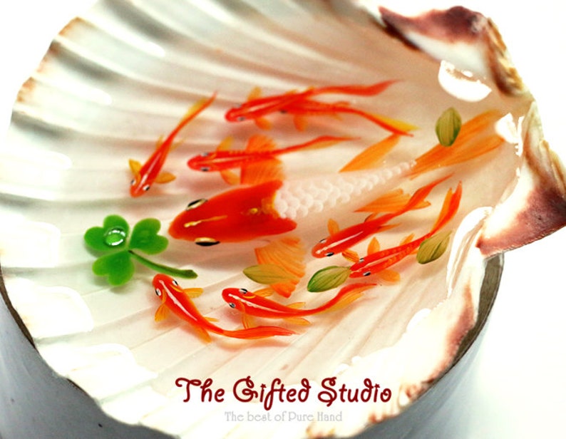 Puede incluir: Una concha decorativa con peces dorados en miniatura de color naranja y blanco nadando en su interior. Tambi&eacute;n hay un tr&eacute;bol verde de cuatro hojas. El texto "The Gifted Studio" est&aacute; en la parte inferior.