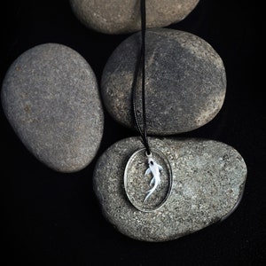 May include: A silver pendant necklace with a white fish encased in resin. The necklace is displayed on a black background with gray stones.