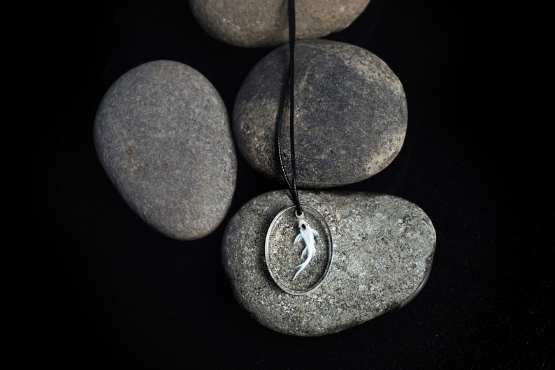 May include: A silver pendant necklace with a white fish encased in resin. The necklace is displayed on a black background with gray stones.