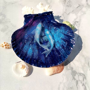 May include: A blue and purple seashell with a galaxy-like design, decorated with two blue and white fish. The seashell is surrounded by smaller seashells.