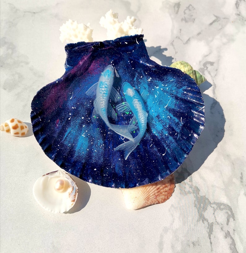 May include: A blue and purple seashell with a galaxy-like design, decorated with two blue and white fish. The seashell is surrounded by smaller seashells.