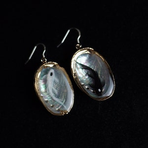May include: A pair of earrings with a gold setting and a mother-of-pearl shell design. One earring features a white fish, while the other features a black fish.