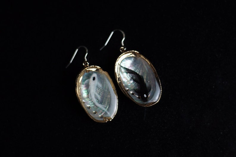 May include: A pair of earrings with a gold setting and a mother-of-pearl shell design. One earring features a white fish, while the other features a black fish.
