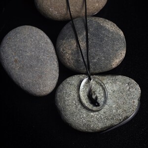 May include: A black fish pendant necklace with a clear resin oval charm. The necklace is on a black cord and is displayed on gray stones.