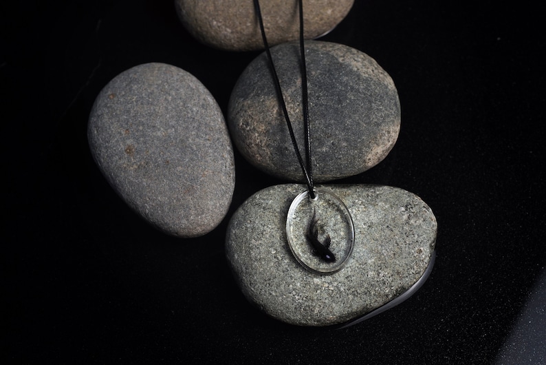 May include: A black fish pendant necklace with a clear resin oval charm. The necklace is on a black cord and is displayed on gray stones.