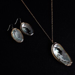 May include: A set of jewelry featuring a necklace and earrings with a mother-of-pearl shell pendant and earrings. The shell is iridescent and has a black and white fish painted inside. The necklace and earrings are gold-toned.