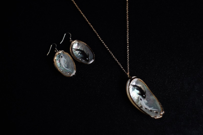 May include: A set of jewelry featuring a necklace and earrings with a mother-of-pearl shell pendant and earrings. The shell is iridescent and has a black and white fish painted inside. The necklace and earrings are gold-toned.