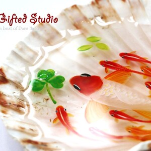 Puede incluir: Una pieza de arte en concha marina con un pez grande blanco y rojo con peces m&aacute;s peque&ntilde;os de color naranja nadando en resina clara y acuosa. Tambi&eacute;n hay un tr&eacute;bol verde y hojas. El texto "The Gifted Studio" es visible.