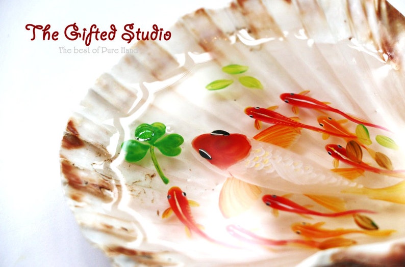 Puede incluir: Una pieza de arte en concha marina con un pez grande blanco y rojo con peces m&aacute;s peque&ntilde;os de color naranja nadando en resina clara y acuosa. Tambi&eacute;n hay un tr&eacute;bol verde y hojas. El texto "The Gifted Studio" es visible.