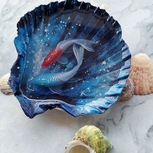 May include: A blue seashell with a resin coating, containing two painted fish. The fish are white with red and blue accents. The seashell is on a white marble surface.