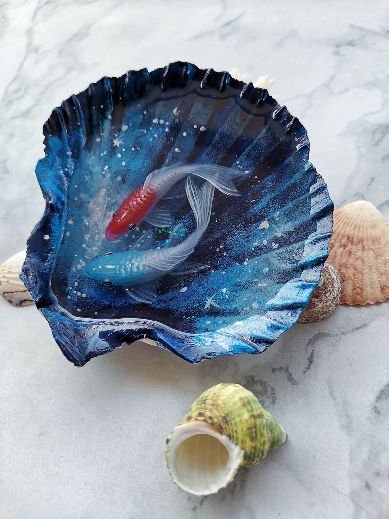 May include: A blue seashell with a resin coating, containing two painted fish. The fish are white with red and blue accents. The seashell is on a white marble surface.