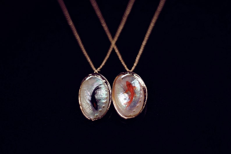 May include: Two gold necklaces with oval-shaped pendants. Each pendant is made of iridescent mother-of-pearl and features a fish inside. One fish is black and the other is orange.