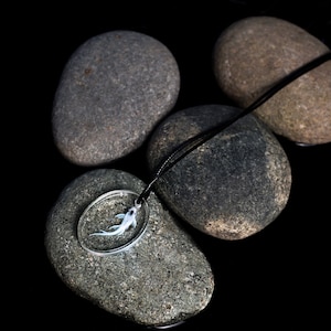May include: A silver necklace with a round pendant containing a small, white fish. The necklace is lying on top of four gray stones against a black background.
