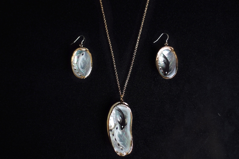May include: A set of abalone shell jewelry featuring a black fish design. The set includes a necklace and earrings with gold chains.