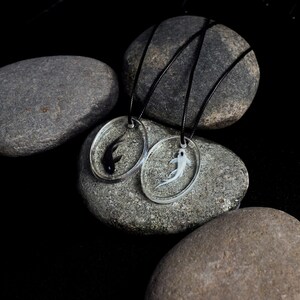 May include: Two necklaces with black cord and round pendants featuring a black and white fish in resin. The necklaces are displayed on gray stones.
