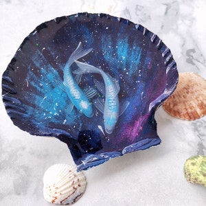 May include: A seashell with a blue and purple galaxy-like design and two white fish painted inside. The seashell is sitting on a white marble surface.