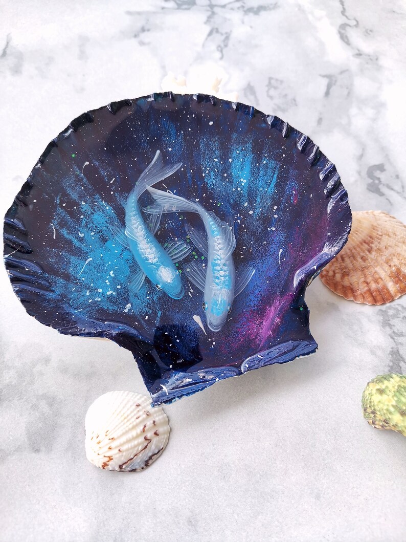 May include: A seashell with a blue and purple galaxy-like design and two white fish painted inside. The seashell is sitting on a white marble surface.