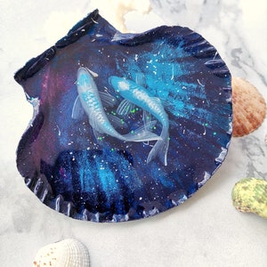 May include: Two white fish with green fins are displayed in a blue and purple seashell with a galaxy design. The seashell is on a white marble surface.