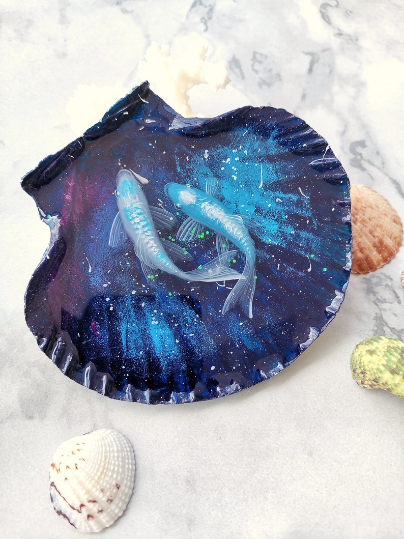 May include: Two white fish with green fins are displayed in a blue and purple seashell with a galaxy design. The seashell is on a white marble surface.