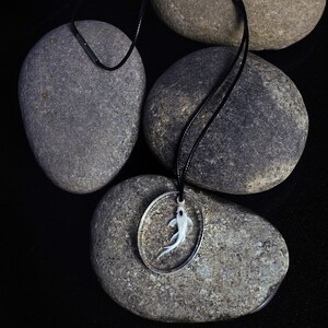 May include: A silver necklace with a round pendant featuring a white fish inside. The necklace is laying on top of four gray stones.