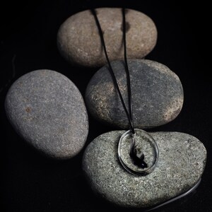 May include: A black cord necklace with a clear glass pendant featuring a black silhouette of a fish. The necklace is displayed on top of four smooth gray stones.