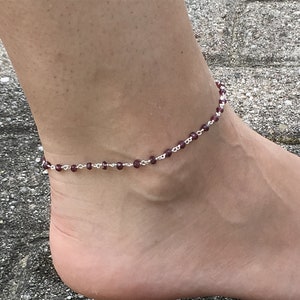 May include: A silver anklet with small, dark red beads. The anklet is on a person's ankle.
