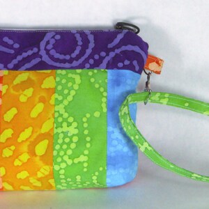 Puede incluir: Una pequeña bolsa con cremallera de colores con un tejido estampado de arcoíris y una correa verde. La bolsa está hecha de un tejido suave de algodón y tiene un cierre de cremallera.