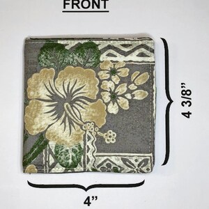 May include: A gray fabric wallet with a floral print featuring green leaves and white hibiscus flowers. The wallet measures 4 inches wide and 4 3/8 inches tall.