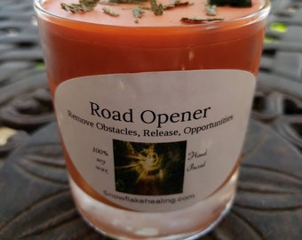 May include: A small, orange-colored candle in a clear glass votive. The candle has a white label with the text "Road Opener" and "Remove Obstacles, Release, Opportunities". The candle is decorated with dried herbs and a small green stone.