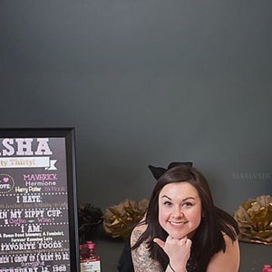 May include: A chalkboard sign with the name Sasha and the text "Dirty Thirty!"  The sign lists personal information such as height, weight, favorite foods, and a birthday date. A woman with long dark hair is smiling and looking at the camera. She is wearing a black top and has a black bow in her hair. The background is a gray wall with black and gold balloons.