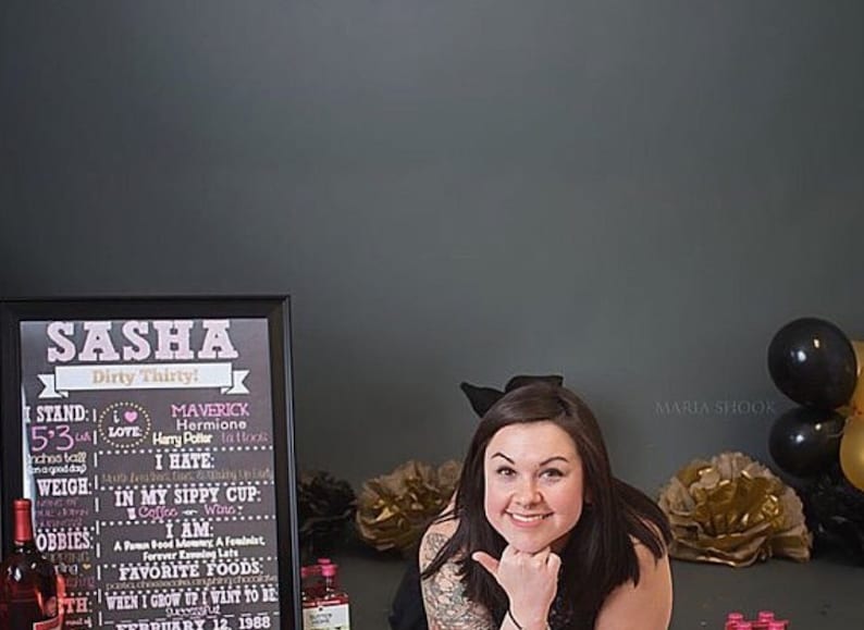 May include: A chalkboard sign with the name Sasha and the text "Dirty Thirty!"  The sign lists personal information such as height, weight, favorite foods, and a birthday date. A woman with long dark hair is smiling and looking at the camera. She is wearing a black top and has a black bow in her hair. The background is a gray wall with black and gold balloons.