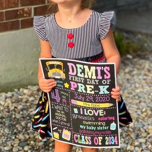 May include: A black and white striped dress with two red buttons and a black skirt featuring a colorful crayon print. The dress is paired with a rectangular chalkboard sign that reads Demi's First Day of Pre-K, July 29, 2020, and Class of 2034.